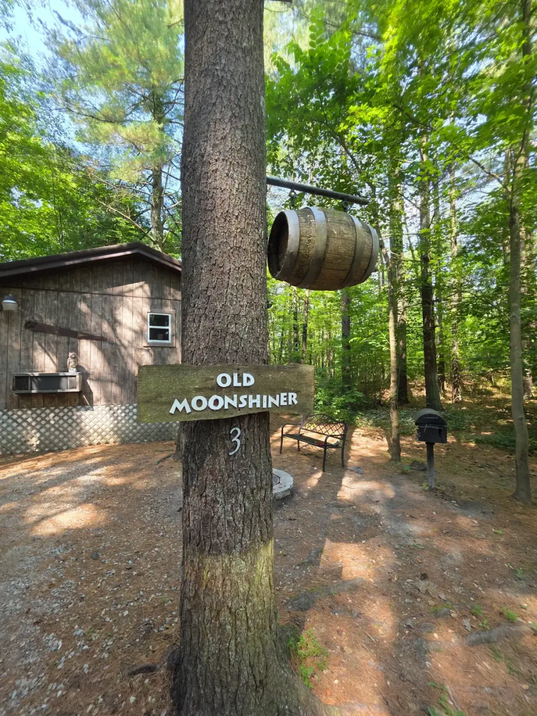 Old Moonshiner name sign on tree with whiskey barrel and wooded pathway to cabin