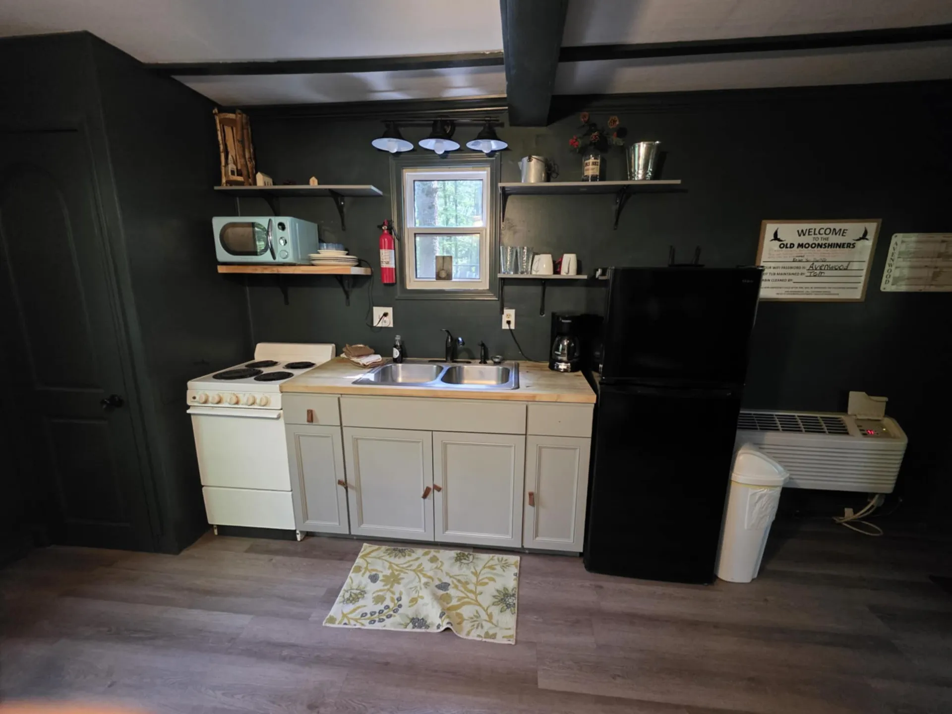 Kitchenette with dark walls, open shelving, compact stove, and Welcome to Old Moonshiner sign
