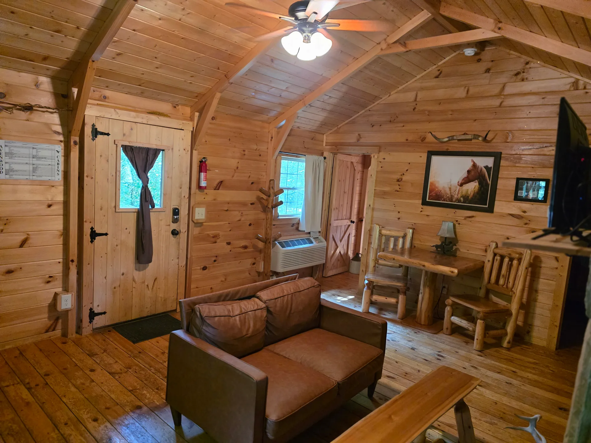 Open-concept living area inside Settlers Roost cabin with leather loveseat, log dining set, and rustic wooden entry door