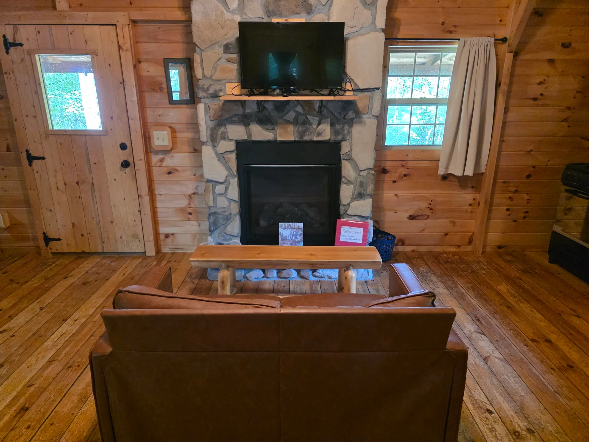 Living room in Settlers Roost cabin with stone fireplace, wall-mounted TV, leather sofa, and rustic coffee table