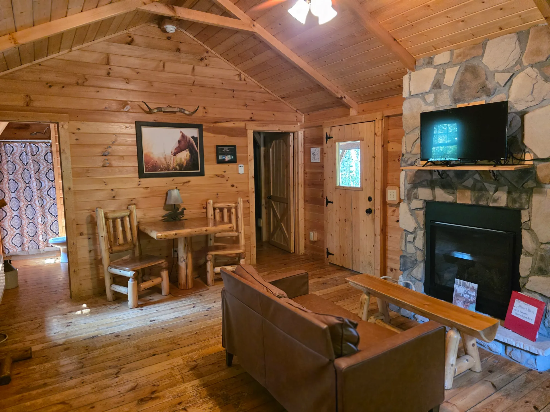Cozy living room inside Settlers Roost cabin with stone fireplace, flat-screen TV, leather sofa, and rustic log dining table